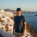A man in a dark blue t-shirt and cap stands in front of the white-washed buildings of Santorini, Greece, overlooking the sea during sunset.