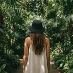 Back view of a woman with long brown hair wearing a black bucket hat and a white strap dress, standing amidst lush green tropical plants in a greenhouse.
