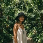A woman wearing a black sun hat and a light beige dress stands amidst lush green tropical plants in a botanical garden.