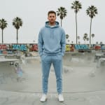 A man in a light blue matching hoodie and sweatpants tracksuit stands in a concrete skatepark with graffiti and palm trees in the background.