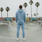 Back view of a man wearing a light blue hoodie and sweatpants tracksuit standing in a concrete skatepark with palm trees and graffiti in the background.