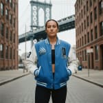 A woman in a blue and white varsity jacket stands on a cobblestone street in Brooklyn with the Manhattan Bridge framed by brick buildings in the background.