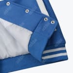 A close up shot of a blue varsity jacket's bottom corner, showing silver snap buttons, a white lining, and a blue ribbed hem with white stripes.