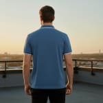Back view of a man in a blue polo shirt with white trim, standing on a rooftop balcony overlooking a city skyline at sunset.
