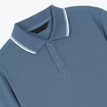 Close-up of a dusty blue pique polo shirt featuring a ribbed collar with white stripes and a two-button placket.