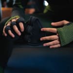 Close-up of a person hands holding and wearing black gym grip pads with finger loops, featuring a green and black striped compression sleeve on the wrist.
