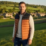A man wearing a navy and orange puffer vest over a cream sweater stands in a grassy field with an English village in the background.