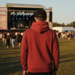 A man from behind wearing a red hooded sweatshirt and a black cap, looking towards a music festival stage.