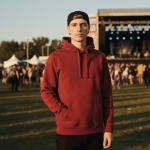A young man wearing a maroon hoodie and a backwards black cap stands in a grassy field at an outdoor music festival during sunset.