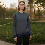 A woman with short brown hair wearing a dark grey crewneck sweatshirt with navy blue cuffs and hem stands in a green park with city buildings in the background.
