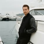A man in a black varsity jacket leans against a white yacht at a marina with other boats in the background.