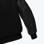 Close-up of a black varsity jacket showing the contrast between the matte body fabric and the textured leather sleeve, with ribbed knit cuffs.