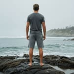 Rear view of a man in a grey t-shirt and shorts standing barefoot on wet rocks, looking out at the ocean waves on a cloudy day.