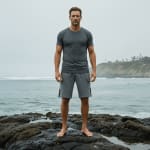 A fit man in a gray t-shirt and gray shorts stands barefoot on dark rocks by the ocean on a cloudy day.