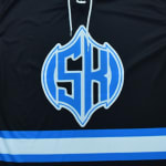 A close-up of a black jersey with a blue and white SK logo in a shield shape and horizontal stripes at the bottom.