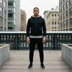 A man standing on a wooden walkway wearing a black hoodie and black sweatpants with city buildings in the background.