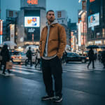 A man in a brown jacket, grey hoodie, and black cargo pants stands on a wet city street in Tokyo at night, with neon billboards and pedestrians in the background.