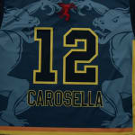 The back of a navy blue sports jersey featuring the number 12 and the name CAROSELLA in black with yellow outlines, flanked by two light blue dragon designs.