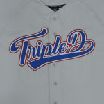 A close-up of a light grey baseball jersey with Triple D written in blue cursive script with a red and white outline across the chest.