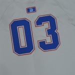 Close-up of a light gray jersey showing the number 03 in blue with red and white borders, topped by a small blue and red logo with a letter D.