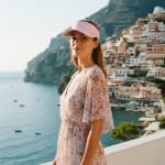 A woman in a pink floral dress and pink visor stands on a balcony overlooking the coastal town of Positano, Italy, with the blue sea and boats in the background.