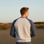 Back view of a man wearing a grey and blue raglan sweatshirt standing in a sandy outdoor field at sunset.