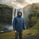 A man in a blue hooded waterproof jacket stands before a large, powerful waterfall surrounded by green mossy cliffs.