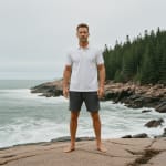 A man standing barefoot on a rocky shore wearing a white polo shirt and dark grey shorts, with crashing ocean waves and a pine forest in the background under an overcast sky.