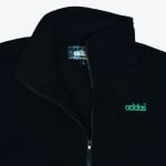 Close-up of a black fleece jacket with a high collar, a zipper, and a green embroidered addos logo on the chest.