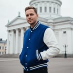 A man wearing a blue and white varsity jacket stands in front of a large white neoclassical cathedral with a dome.