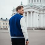 A man from behind wearing a blue varsity jacket with white sleeves, standing in an open square with a large white neoclassical cathedral in the background.
