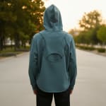 Rear view of a person wearing a teal hoodie with a unique geometric cutout design on the back, standing on an outdoor path at sunset.