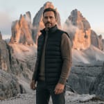 A bearded man wearing a black puffer vest and brown sweater stands against a backdrop of towering, sunlit mountain peaks in the Dolomites.