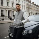 A man in a grey and white letterman jacket and black pants leans against the front of a dark blue Bentley on a city street.