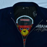 Close-up of a navy blue Urbano All Stars jacket, partially unzipped to show a black lining with a cartoon bear graphic wearing a cap.