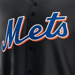 Close-up of a black New York Mets baseball jersey featuring the blue Mets script logo outlined in white and orange.