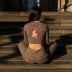 Back view of a woman sitting cross-legged on stone steps, wearing a brown long-sleeved crop top with a tiger and red sun graphic on the back.