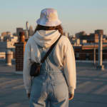 Back view of a person in denim overalls, a cream hoodie, and a tie-dye bucket hat standing on a rooftop looking at a city skyline at sunset.
