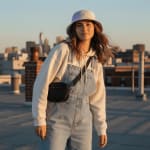 A young woman wearing a bucket hat and denim overalls stands on a rooftop with a blurred city skyline behind her during sunset.