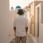 Back view of a man in a white linen shirt and bucket hat standing in a narrow white alleyway in Santorini with a blue-domed church in the background.