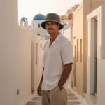 A man wearing a white linen shirt and a green bucket hat stands in a narrow white alleyway in Santorini with a blue-domed church in the background.