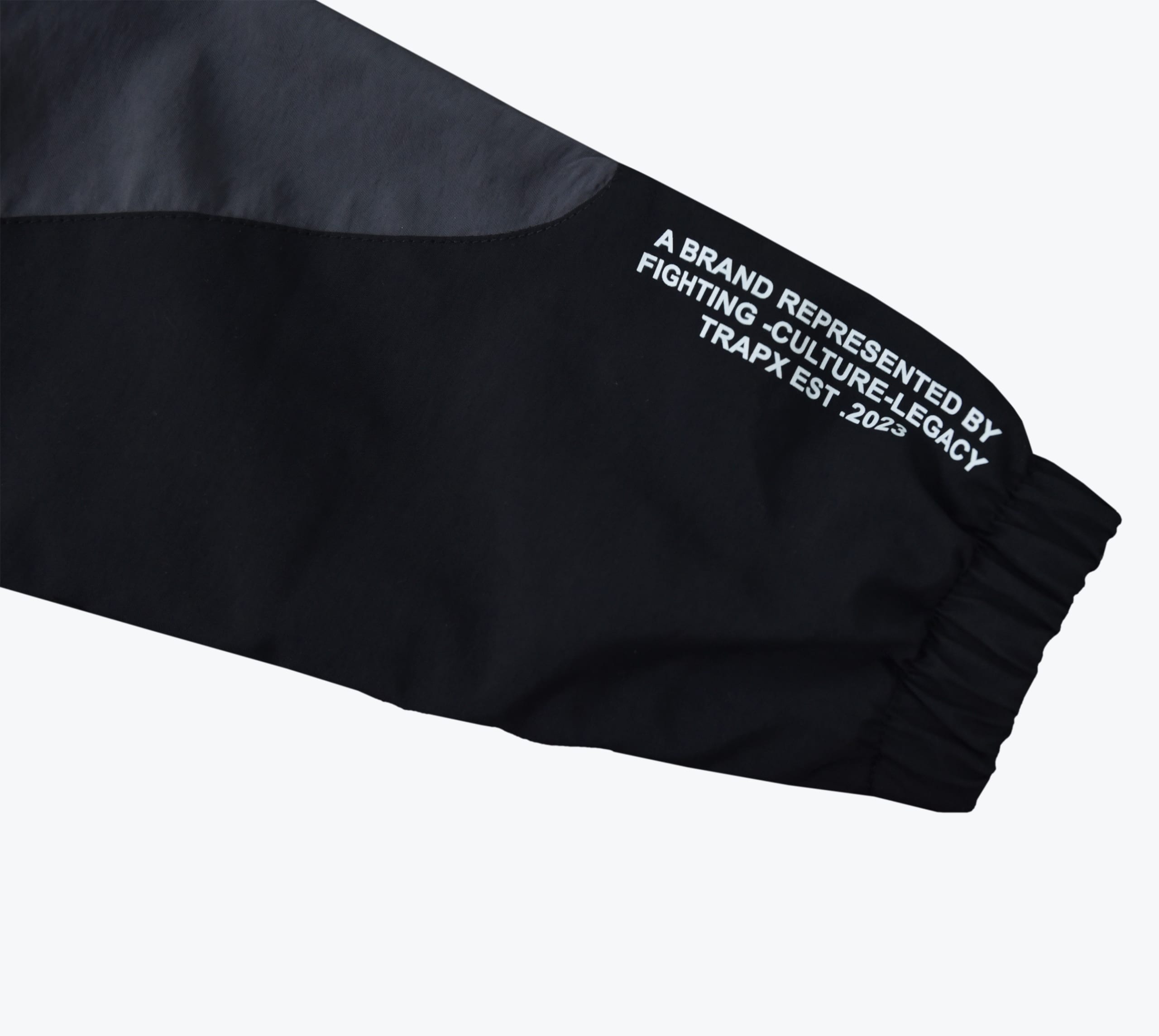 Close-up of a black and grey jacket sleeve with white text reading A BRAND REPRESENTED BY FIGHTING -CULTURE-LEGACY TRAPX EST .2023 near the elastic cuff.