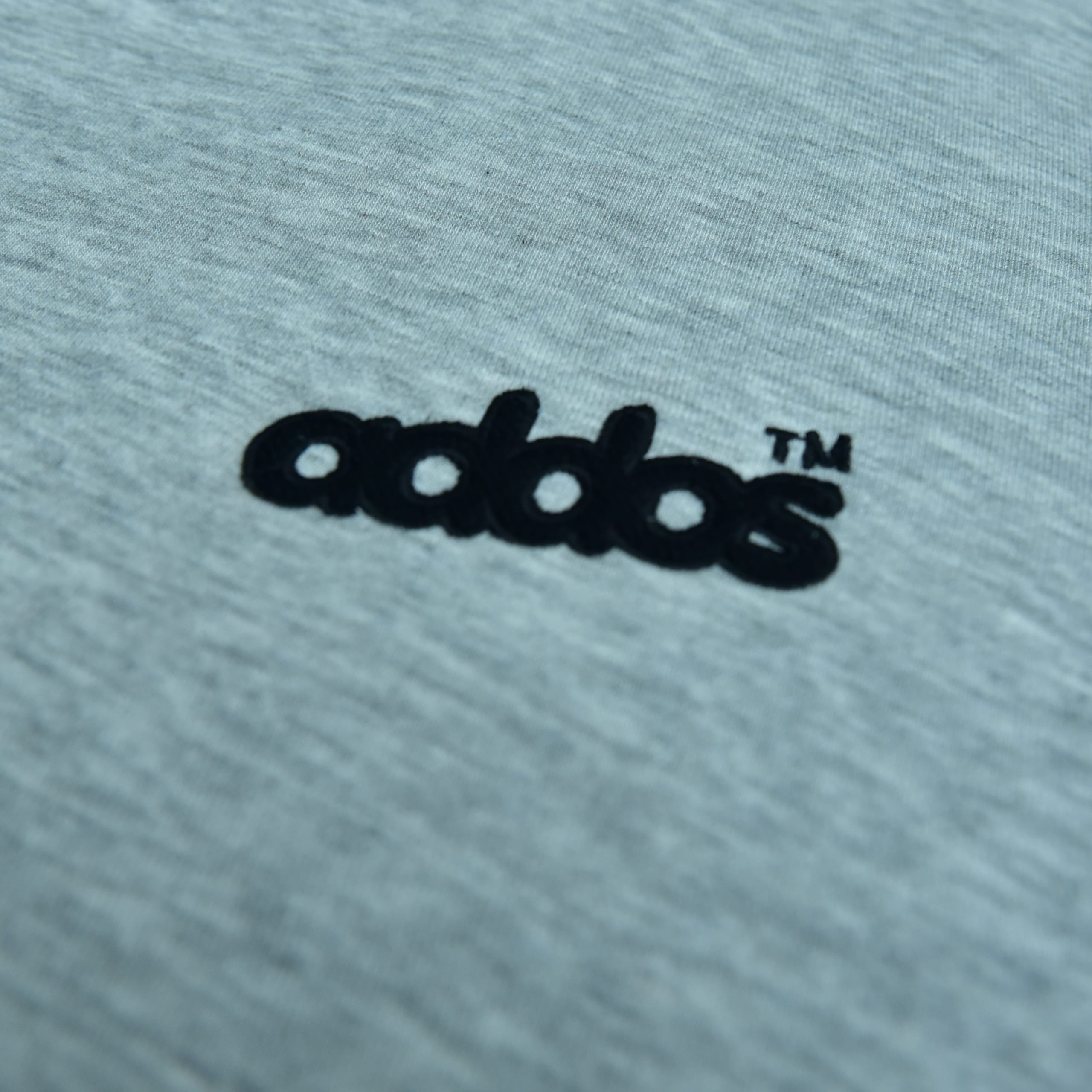 Close-up of a grey heather fabric with a black embroidered logo reading addos with a TM trademark symbol.