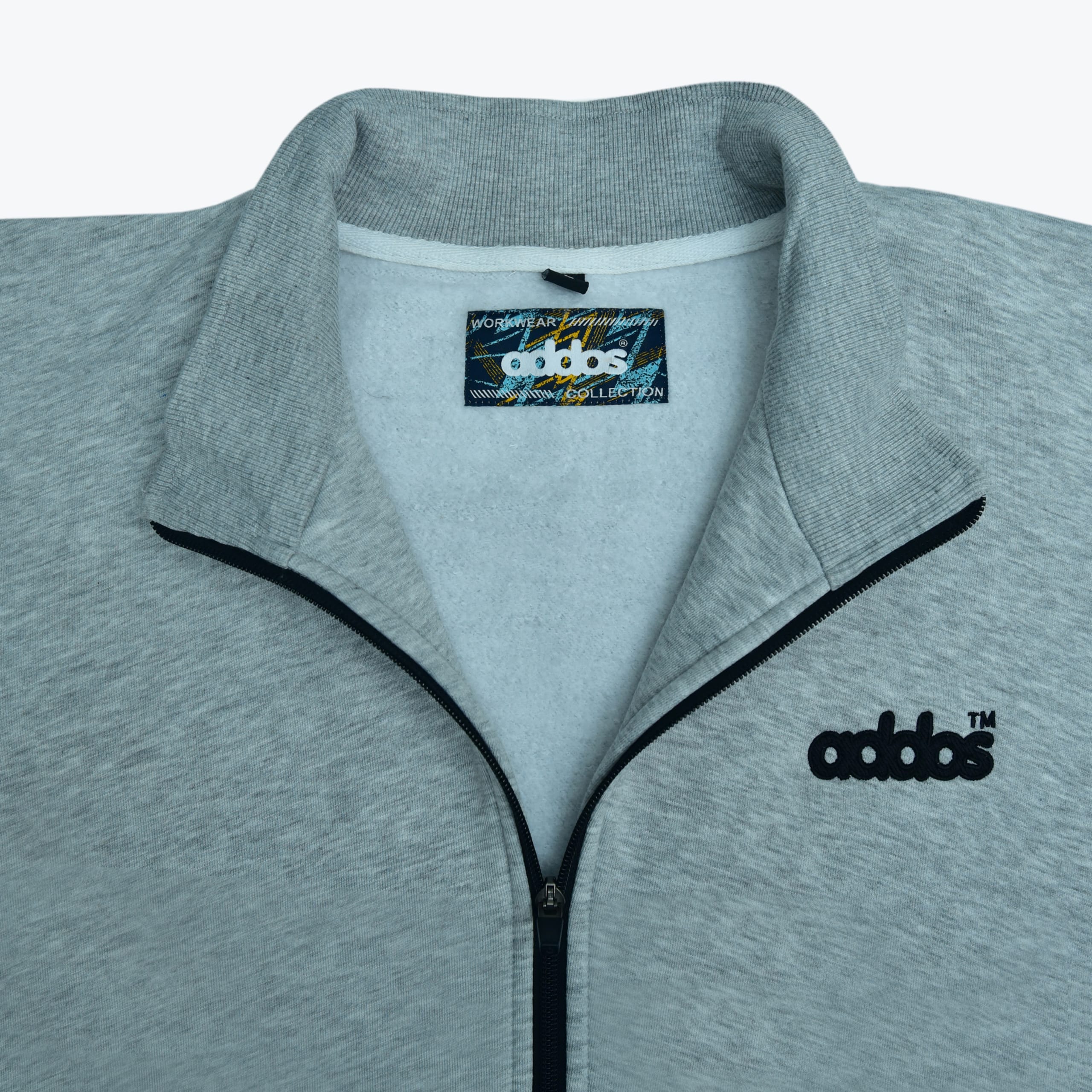 Close-up of a grey zip-up jacket with a ribbed collar, black zipper, and addos embroidered in black on the left chest.