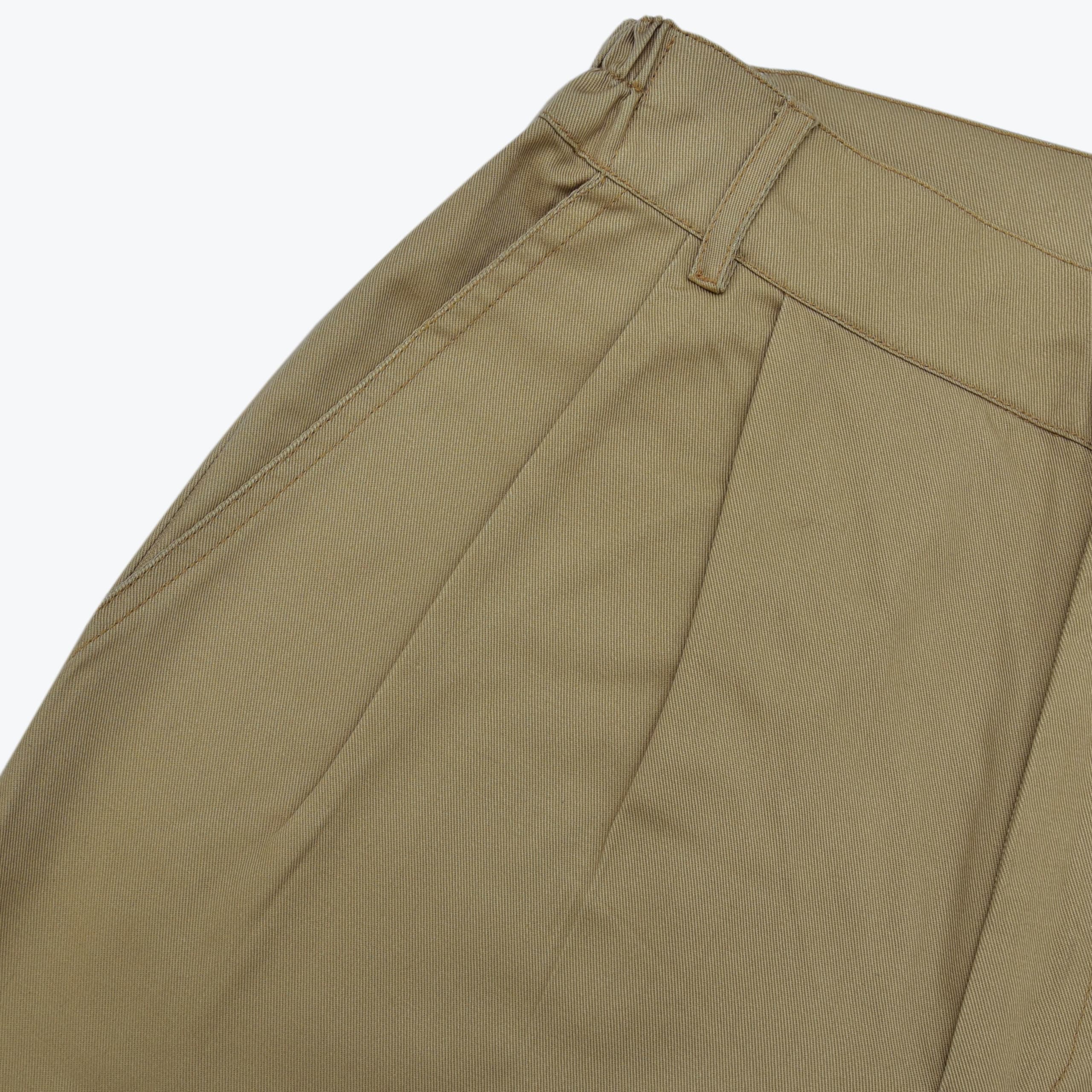 Close-up view of the waistband, pleats, belt loops, and side pocket of a pair of tan khaki trousers against a white background.