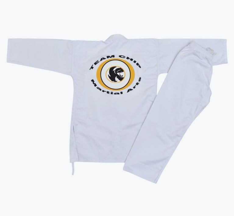 A flat lay of a white martial arts uniform jacket and pants, featuring a circular Team Chip Martial Arts logo on the back of the jacket.