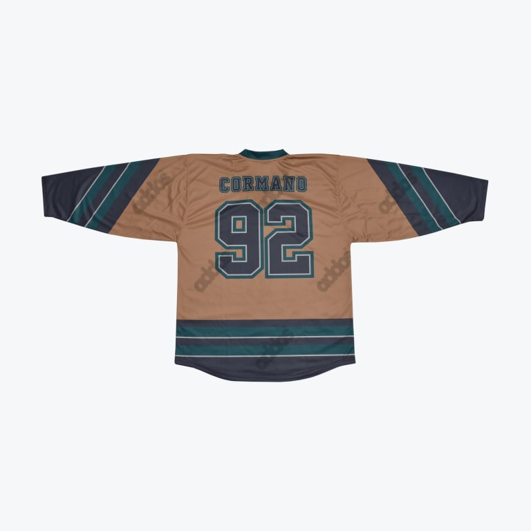 The back of a tan hockey jersey with CORMANO and 92 printed in dark teal and navy block letters, featuring striped sleeves and hem.
