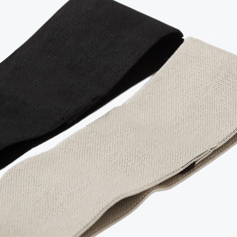 Close-up of a black elastic band and a beige elastic band laid diagonally on a white background, showing their woven texture.