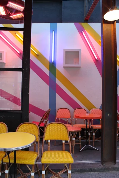 Yellow rattan chairs in the foreground on Le Tournesol's terrace. Behind them, orange chairs and colorful neon lights on the wall. A perfect spot to learn French in Paris