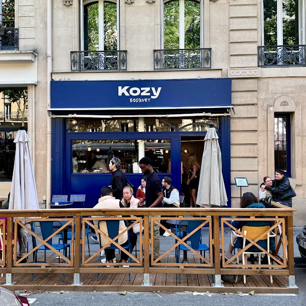 Vue extérieure du café Kozy Bosquet, avec sa devanture bleue, ses tables et des chaises en métal bleu, disposées sur une terrasse.